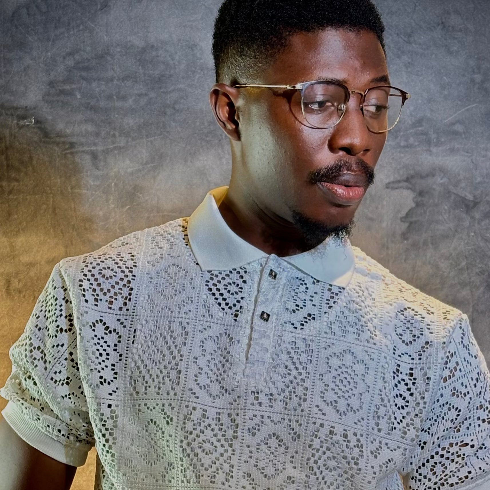 A man wearing a white short-sleeve polo shirt with an intricate openwork crochet or lace pattern, styled with gold-rimmed glasses and a watch, set against a textured gray studio background.